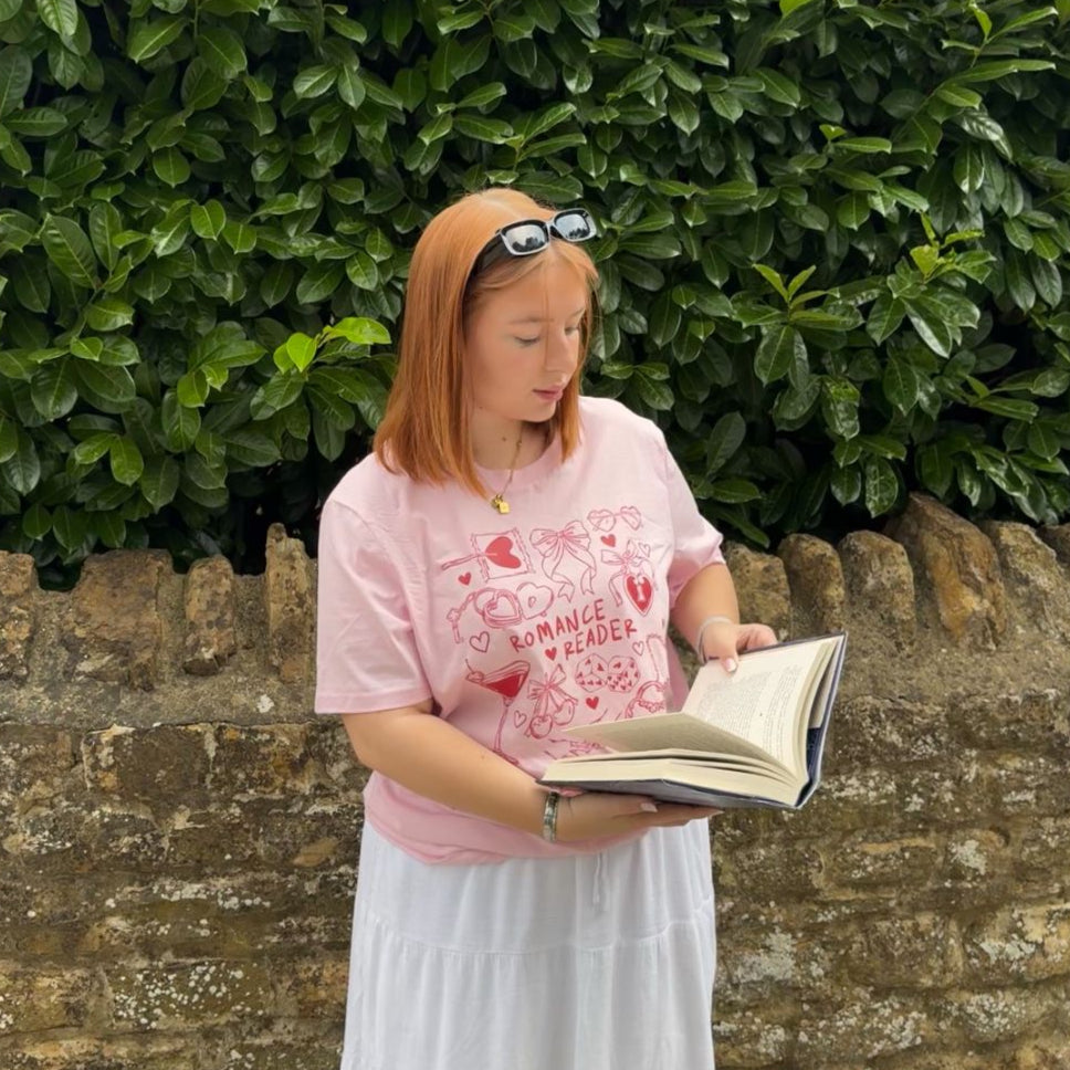 Woman reading a book outdoors against a green hedge and stone wall.