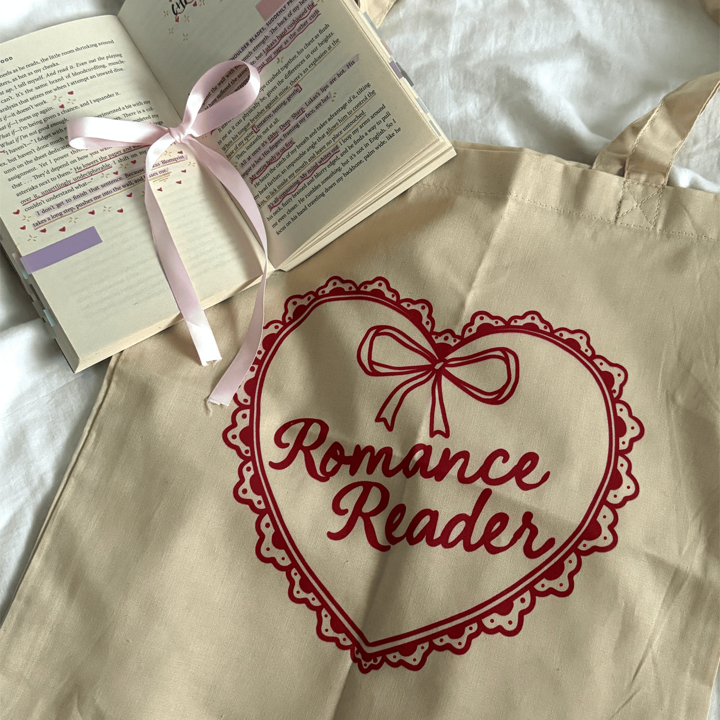 Beige tote bag with 'Romance Reader' design, open book with pink ribbon on a white surface