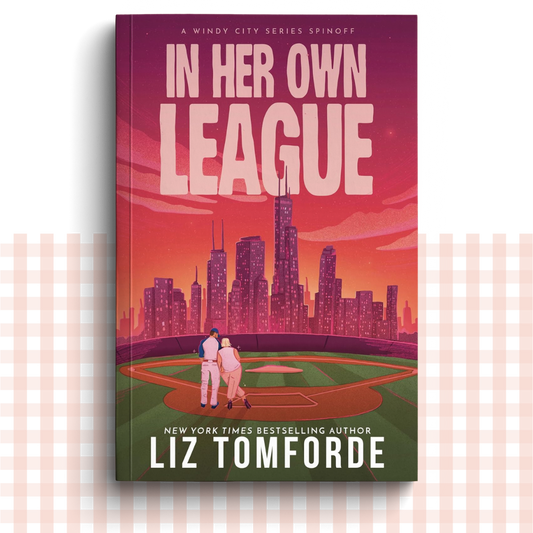 Paperback book cover of 'In Her Own League' by Liz Tomforde with a city skyline and couple on a baseball field.
