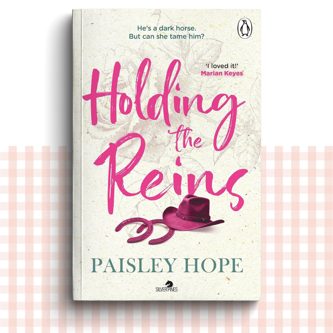 Paperback book cover of 'Holding the Reins' by Paisley Hope with a pink and cream design, and a pink cowboy hat and horseshoes.