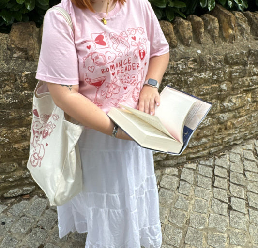 Person wearing a pink 'Romance Reader' t-shirt holding an open book against a stone wall.