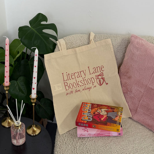 A beige canvas tote bag printed with 'Literary Lane Bookshop' in red lettering, styled on a light beige sofa with a pink pillow, a book, candles, and green leaves.