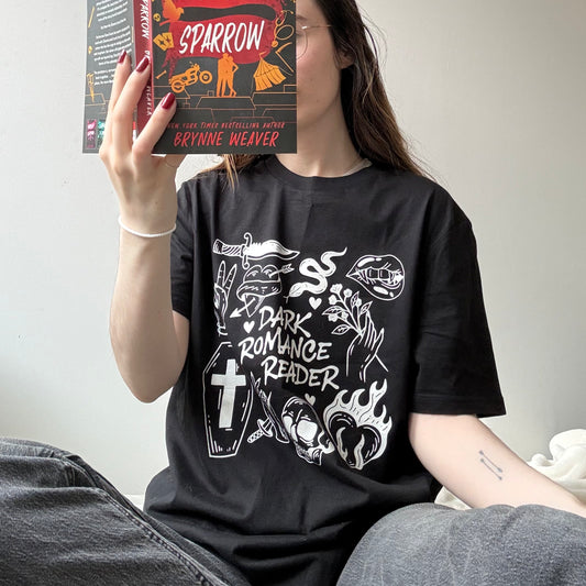 Person wearing a black t-shirt with white graphics and text, holding a book titled 'Scythe and Sparrow' by Brynne Weaver.