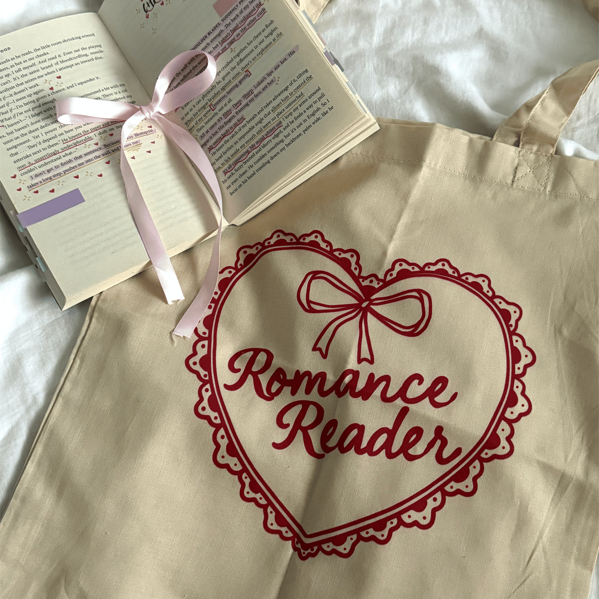 Beige tote bag with 'Romance Reader' design, open book with pink ribbon on a white surface